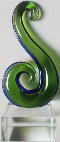 A piece of glass art in the shape of a koru, with green and blue colors, displayed on a clear stand.